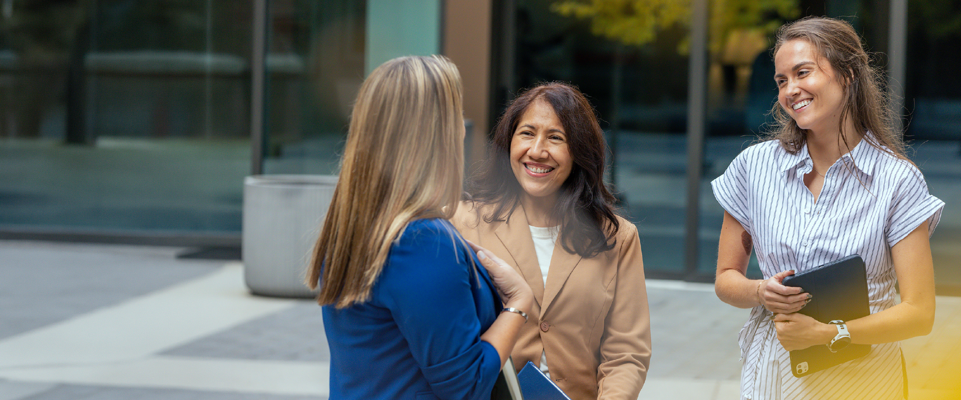 Three of PTC Therapeutics' Patient Engagement team chatting and smiling. One has long light brown hair and is wearing a blue blazer, another, long brown hair, smiling and wearing a camel colored blazer, the third with long brown hair, wearing a pinstriped dress